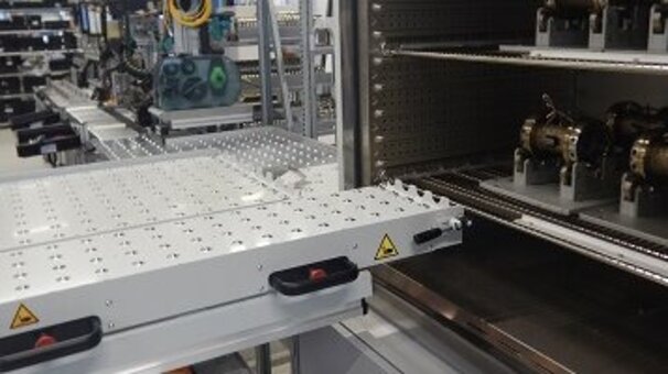 Industrial production facility with an open test cabinet in the foreground containing workpieces. A lifting platform is located in front of the test cabinet.