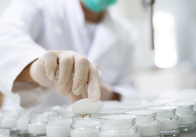 Proben kosmetischer Creme in einem Labor. Im Hintergrund ein Labormitarbeiter in mit Handschuhen und Mundschutz.