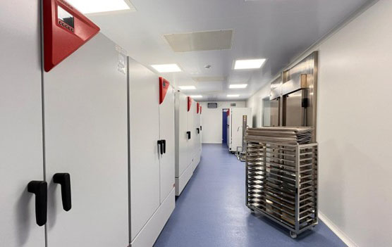 Corridor with several BINDER climate chambers with red triangular controls, blue floor, and stainless steel trolley with trays in a laboratory or testing facility room.