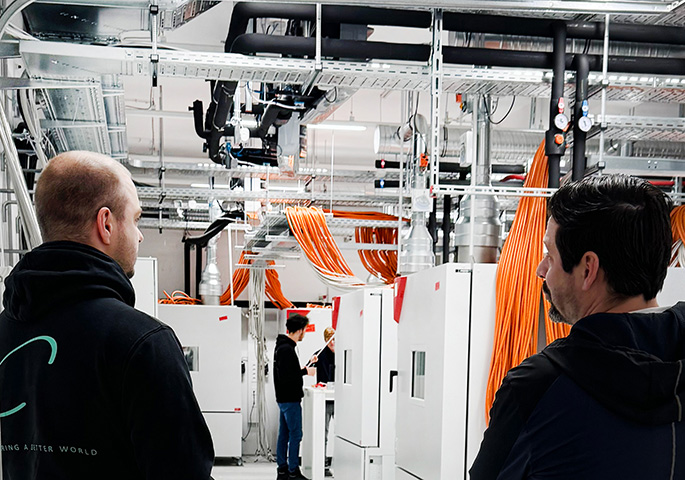 People are standing in a battery test laboratory with BINDER battery test chambers. Numerous orange-colored cable harnesses, silver ventilation pipes, and lines run across the ceiling. Two people in the foreground are looking at the system, while others in the background are working on the units.